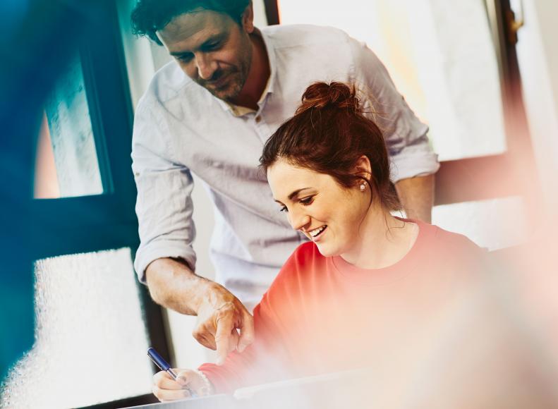 man standing next to a woman, pointing to a paper on her desk